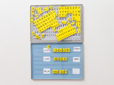 Eine flache, aufklappbare Metallbox, die kleine Magnetblättchen enthält. Alle Magnetblättchen sind mit Buchstaben und Symbolen bedruckt, womit auf dem aufgeklappten Deckel der Box Wörter gelegt werden können.
