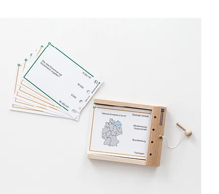 Rechteckige, beschriftete Pappkarten mit Löchern am rechten Rand liegen aufgefechert auf einem Tisch. Daneben befindet sich ein offener Holzkasten, auf dessen Rand kleine Löcher gebohrt sind. Am Holzkasten ist eine Holznadel mit einem Band befestigt.