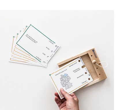 Rechteckige, beschriftete Pappkarten mit Löchern am rechten Rand liegen neben einem offenen Holzkasten. Eine Hand legt eine der Pappkarten in den Holzkasten. 