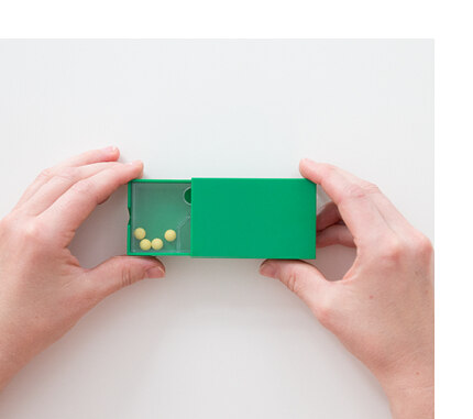 Die grüne Schüttelbox wird von zwei Händen bis zur Hälfte ausgezogen und es sind 4 Kügelchen auf der linken Seite des Inlays sichtbar.