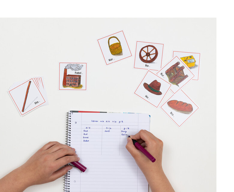 Qudratische Karten, auf denen Gegenstände und einzelne Wortsilben zu sehen sind, liegen ungeordnet nebeneinander. Darunter liegt ein Schreibblock, auf den jemand Wörter schreibt.