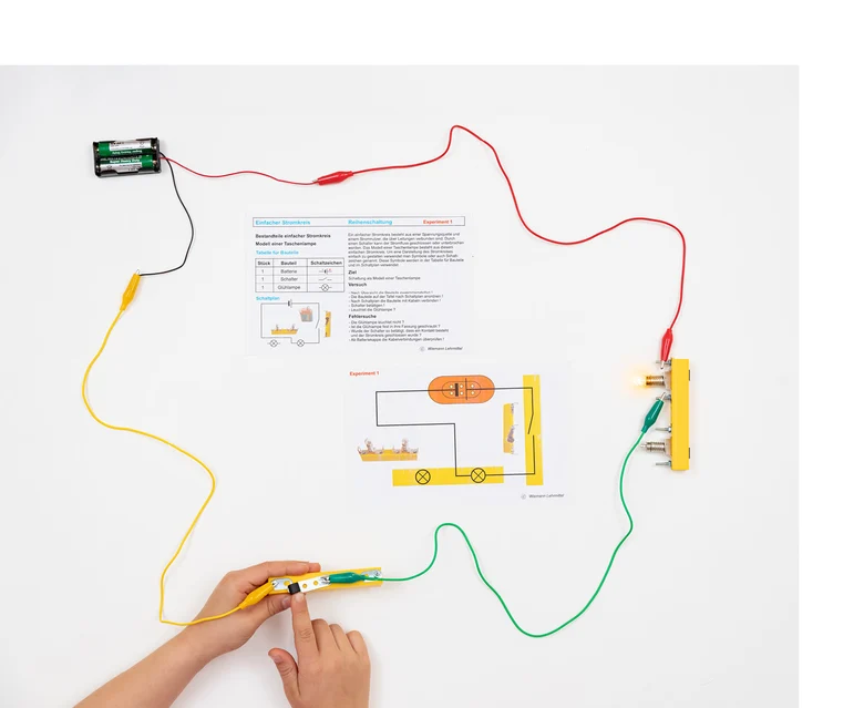 Die bunten Kabel und einzelne Bauteile sind miteinander verbunden. In der Mitte liegt eine Beschreibung des aufgebauten Experiments.