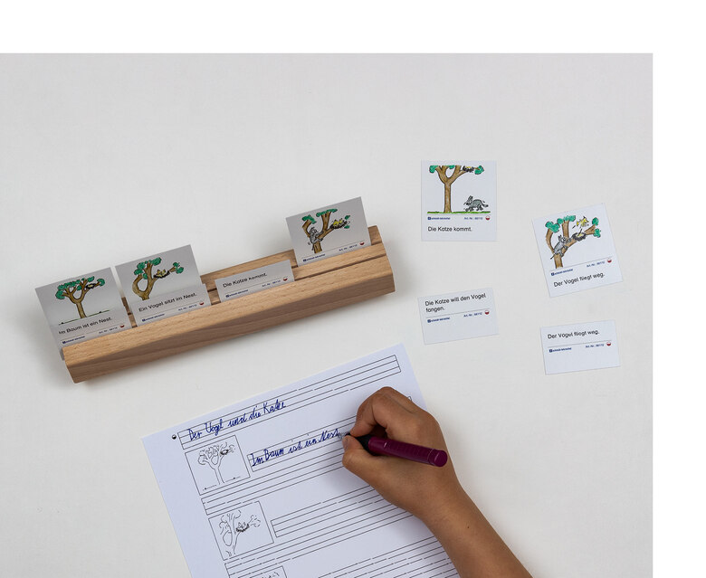 Auf einer Holzleiste stecken Karten mit Bildern und dem dazu passenden Satz. Darunter ein Blatt Papier, auf dem eines der Bilder abgebildet ist und Linien, auf denen der dazu passende Satz geschrieben wird.