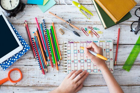 Viele bunte Lern- und Schreibutensilien liegen auf einem Holztisch. Eine Kinderhand schreibt auf einem Schreibblock. 