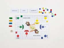 Ein Spielfeld mit verschiedenen Feldern. Daneben Spielfiguren, ein Würfel und Karten mit Wörtern darauf.